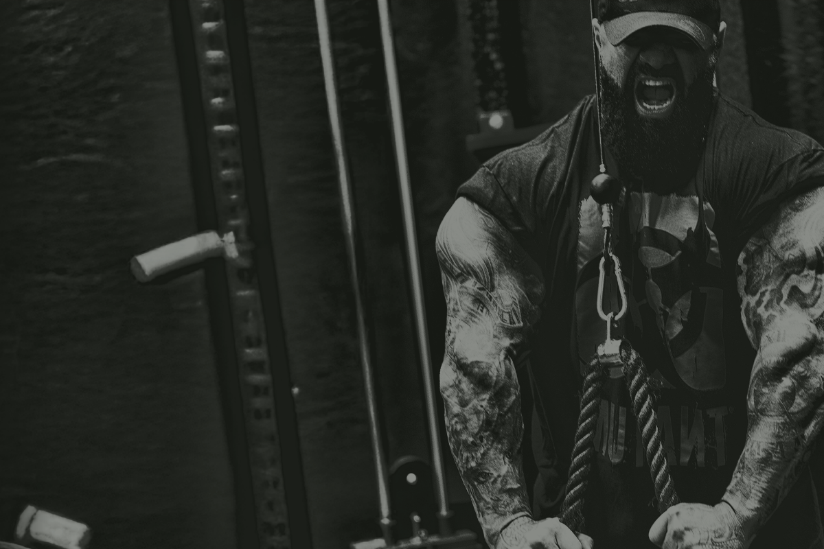 Black-and-white image of a muscular, tattooed man in a cap shouting with effort while performing a cable rope exercise in a gym.