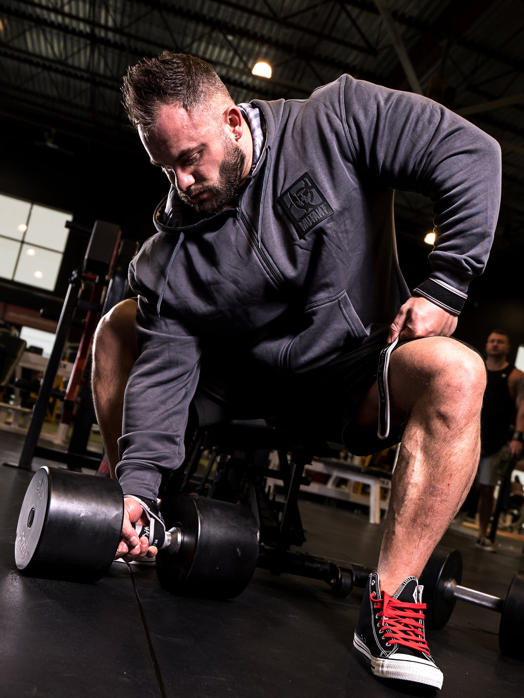 Mutant athlete Dana Baker striking a pose in the gym, wearing the Grey Mutant Patched Zip-Up Gym Hoodie. The hoodie features a black Mutant logo patch on the left chest, two pockets, and a light grey hood lining with a watermarked Mutant logo.