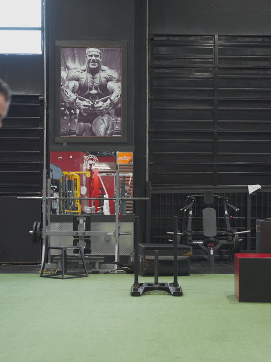 A video of Dana Baker at the gym, posing to showcase both the front and back of the grey 'MUTANT's Truck Month' t-shirt, which features the 'Guts, Glory, Mutant' slogan and the MUTANT logo in black.